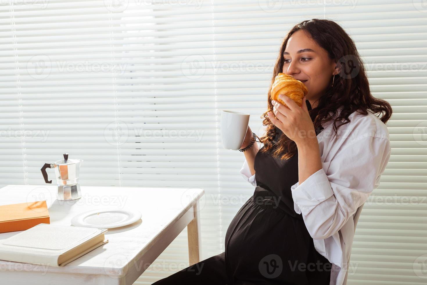 Happy pregnant young beautiful woman eating croissant during morning breakfast. Concept of pleasant morning and positive attitude during pregnancy photo