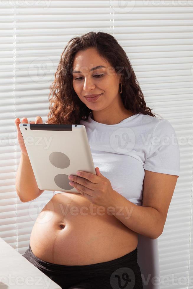 mujer embarazada hispana usando tableta digital mientras desayuna. tecnología, embarazo y licencia de maternidad foto