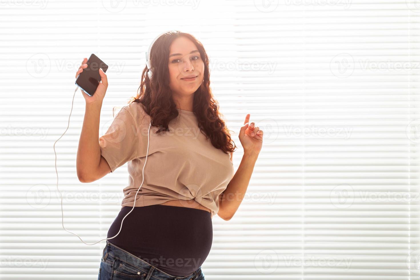 Curly-haired brunette pacified pregnant woman listens to pleasant classical music using smartphone and headphones. Concept of a soothing mood before meeting baby. Copyspace photo