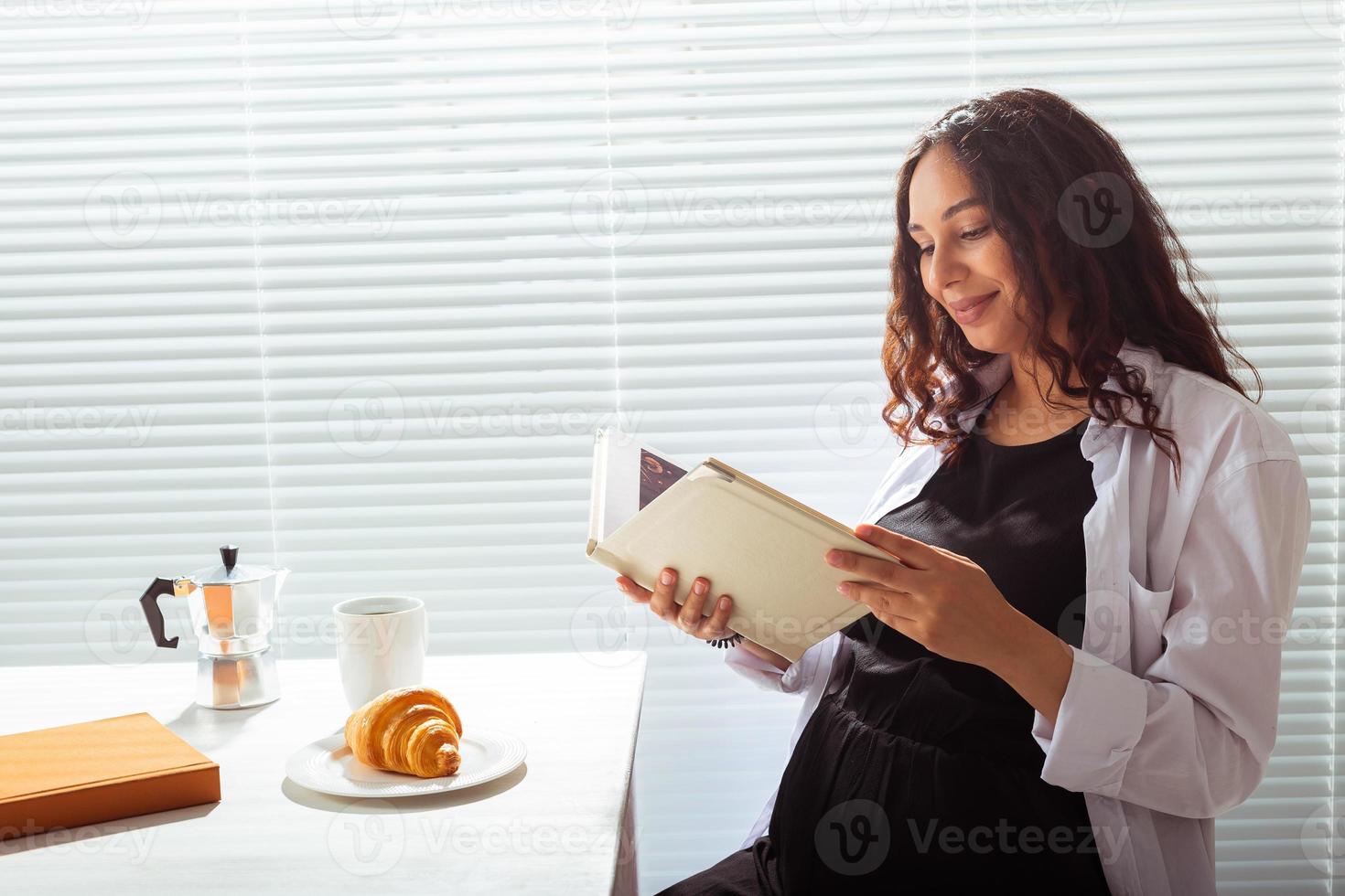 Side view of pregnant woman reading book while having morning breakfast with coffee and croissants on background of blinds. Good morning concept and pleasant lunch break photo