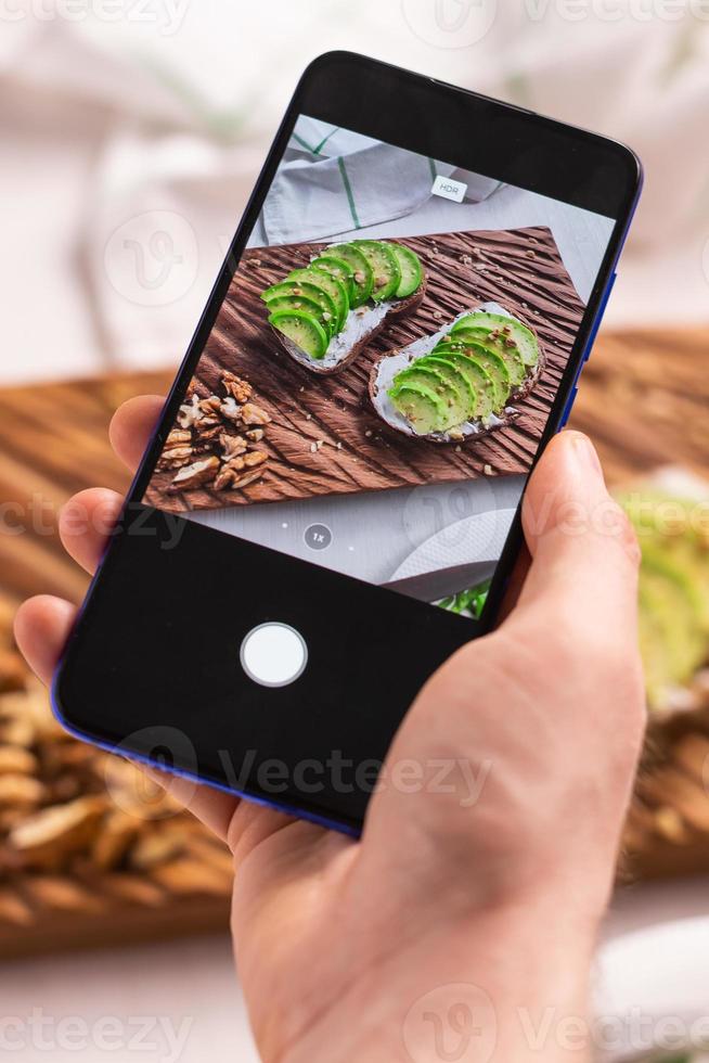 Hands take pictures on smartphone of two beautiful healthy sour cream and avocado sandwiches lying on board on the table. Social media and food concept photo