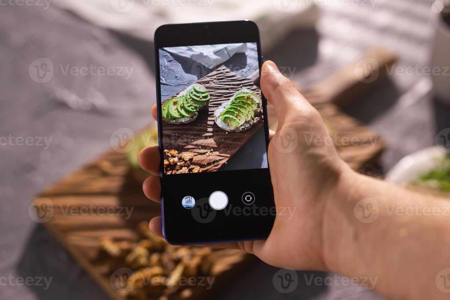 Hands take pictures on smartphone of two beautiful healthy sour cream and avocado sandwiches lying on board on the table. Social media and food concept photo