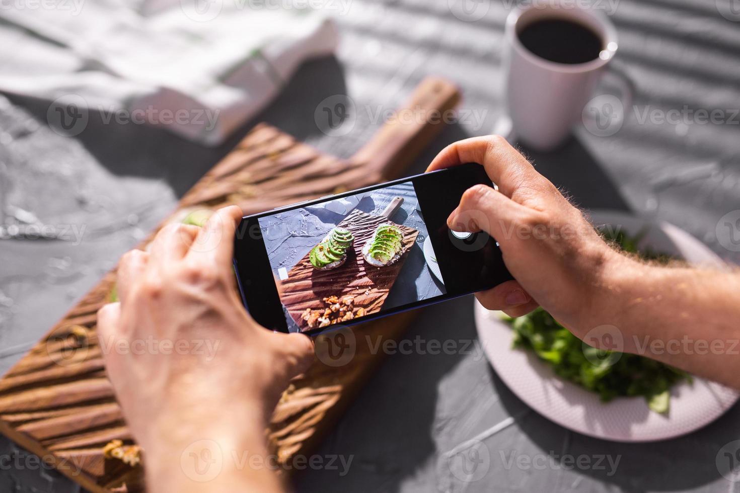 Hands take pictures on smartphone of two beautiful healthy sour cream and avocado sandwiches lying on board on the table. Social media and food concept photo