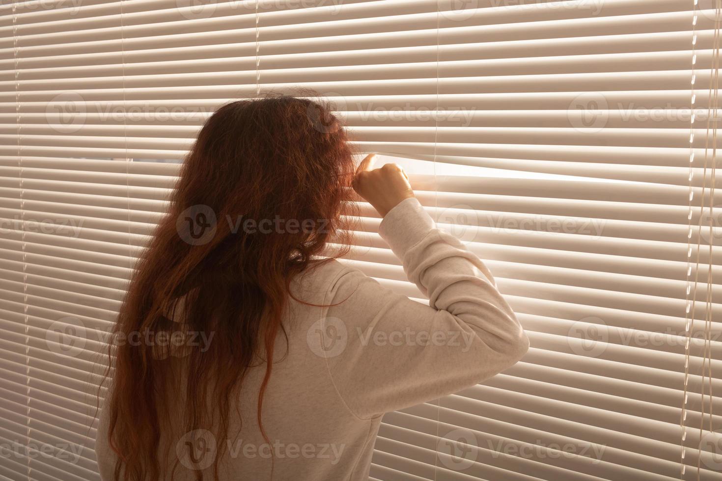 Rear view of beautiful young woman with long hair peeks through hole in the window blinds and looks out the window. Surveillance and curiosity concept photo