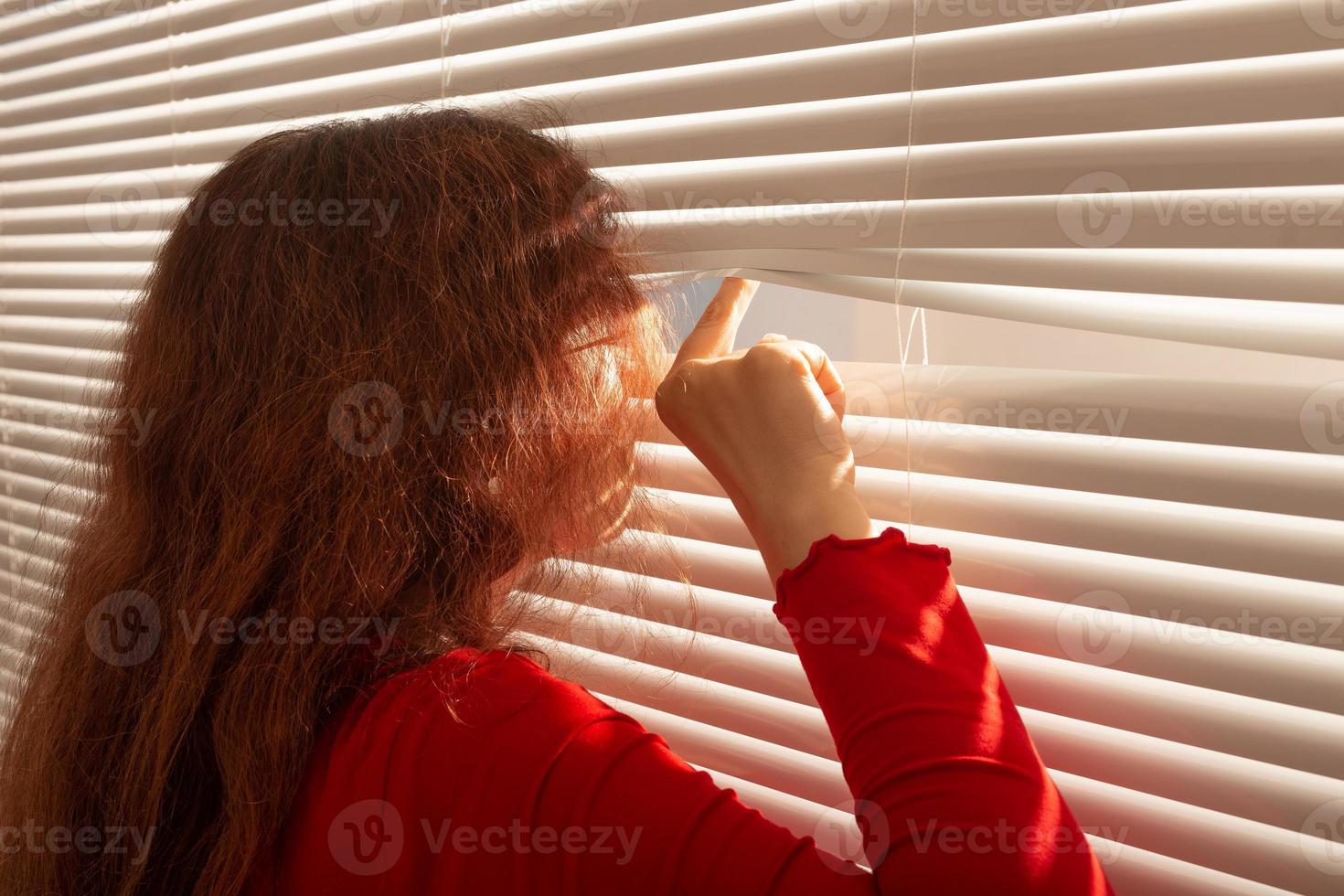 Rear view of beautiful young woman with long hair peeks through hole in the window blinds and looks out the window. Surveillance and curiosity concept photo