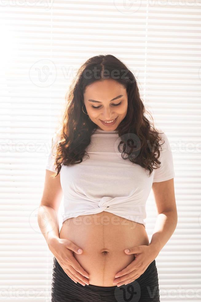 Smiling young beautiful pregnant woman touching her belly and rejoicing. Concept of positive and pleasant feelings while waiting for the baby photo