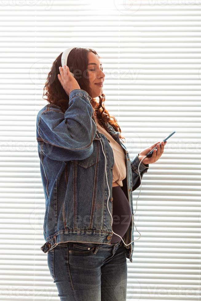 Beautiful young peaceful pregnant woman listens to pleasant classical music using smartphone and headphones. Concept of positive attitude before childbirth photo