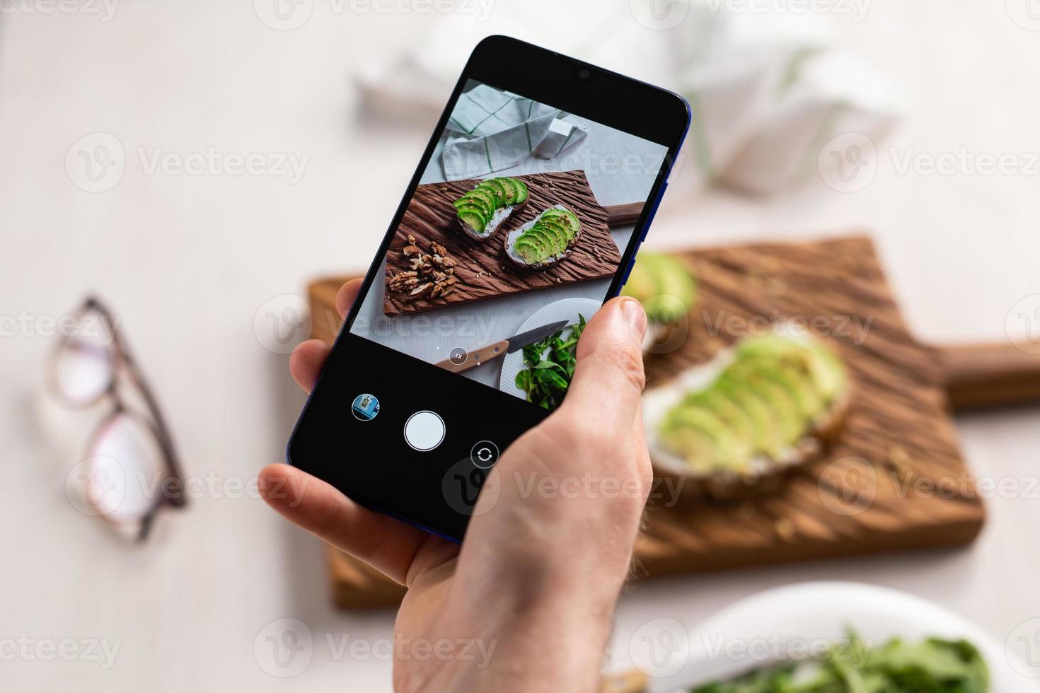 Hands take pictures on smartphone of two beautiful healthy sour cream and avocado sandwiches lying on board on the table. Social media and food concept photo