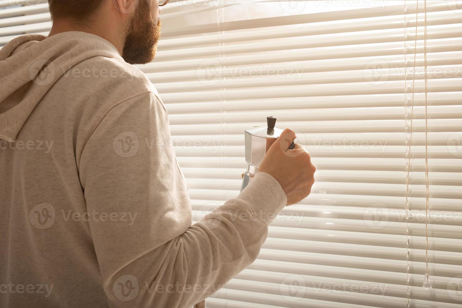 Rear view of young stylish man peeking through hole in window blinds and looking out into street. Concept of enjoying the morning sun and positivity photo