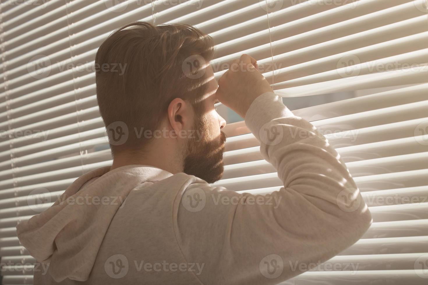 Rear view of young man with beard peeks through hole in the window blinds and looks out into the street. Surveillance and curiosity concept photo