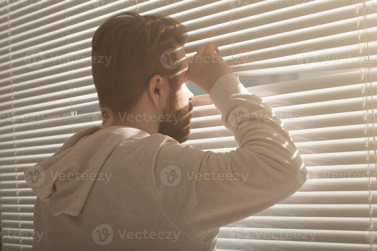 Rear view of young man with beard peeks through hole in the window blinds and looks out into the street. Surveillance and curiosity concept photo