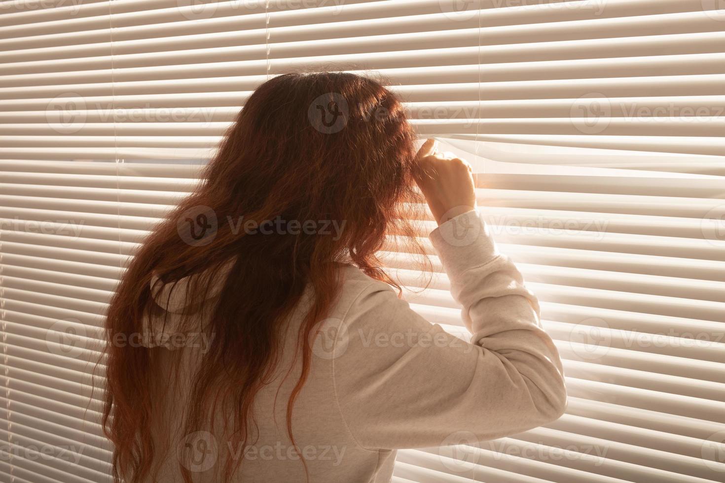 Rear view of beautiful young woman with long hair peeks through hole in the window blinds and looks out the window. Surveillance and curiosity concept photo