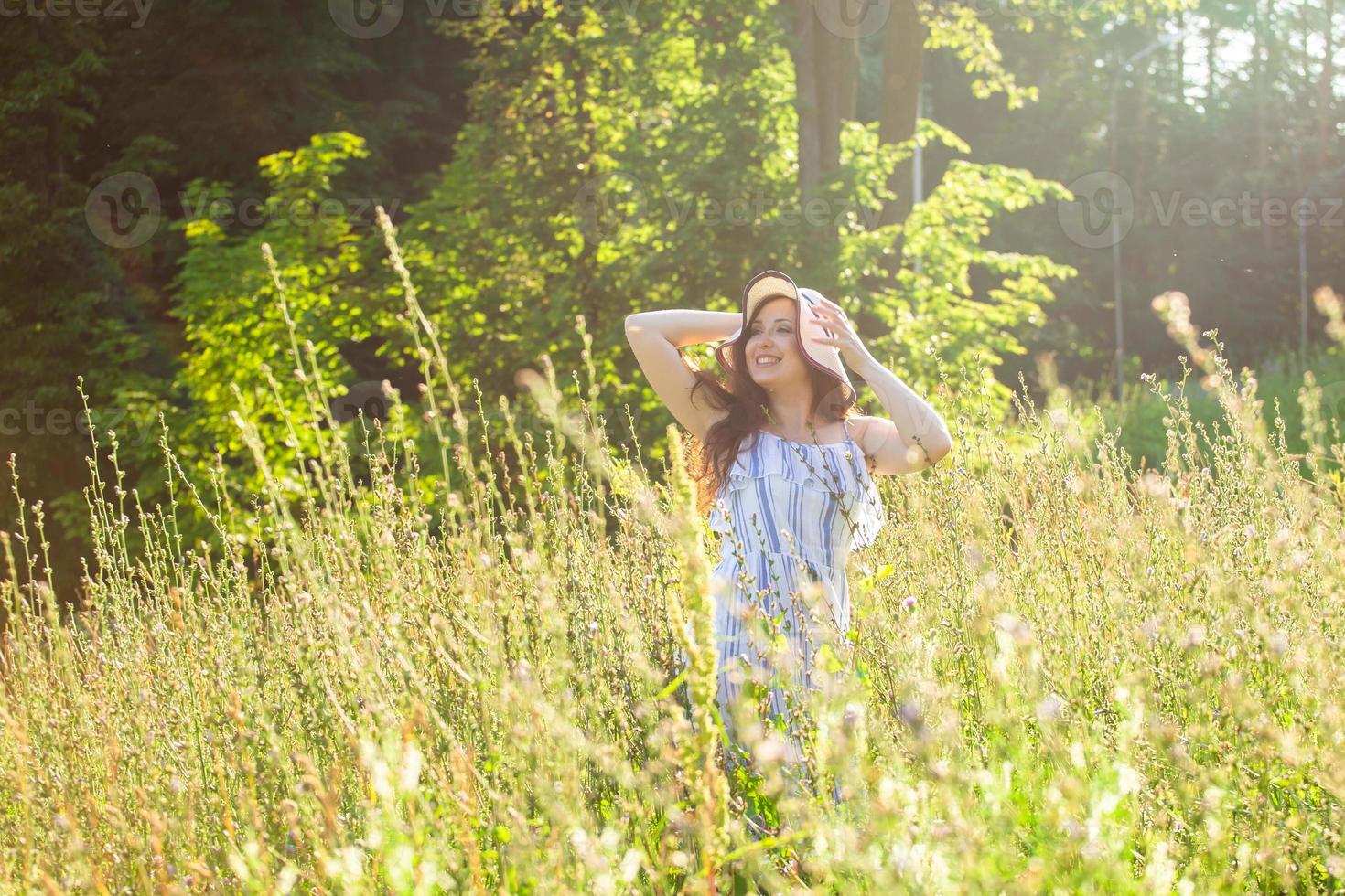 mujer joven feliz con el pelo largo con sombrero y vestido caminando por el bosque de verano en un día soleado. concepto de alegría de verano foto