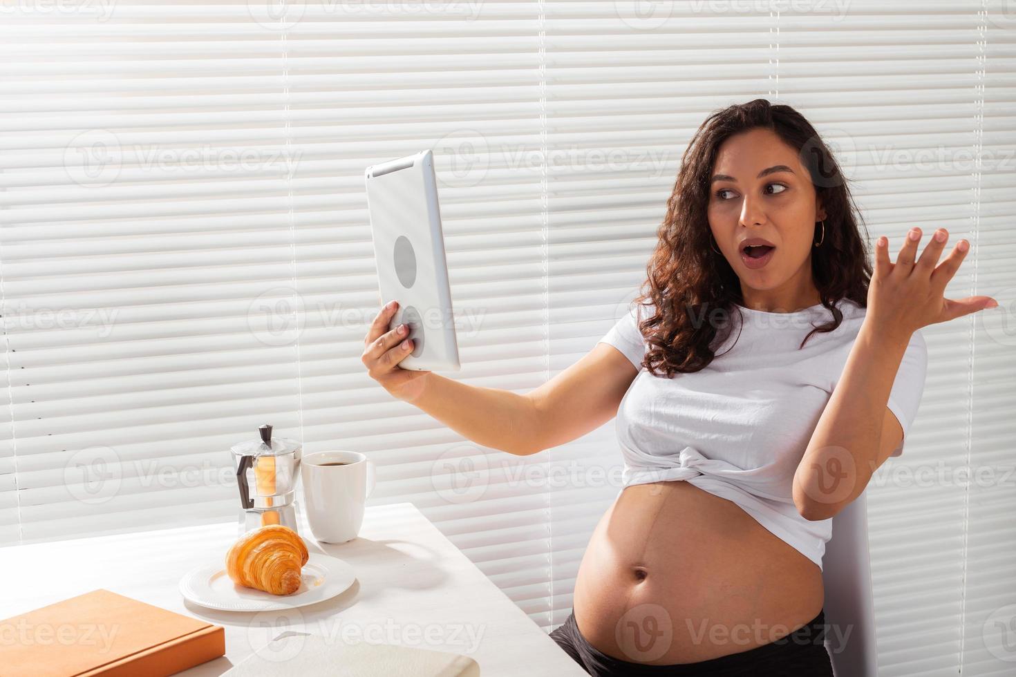 Happy pregnant young beautiful woman talking to mom using video call during morning breakfast. Concept of communication and positive attitude during pregnancy photo