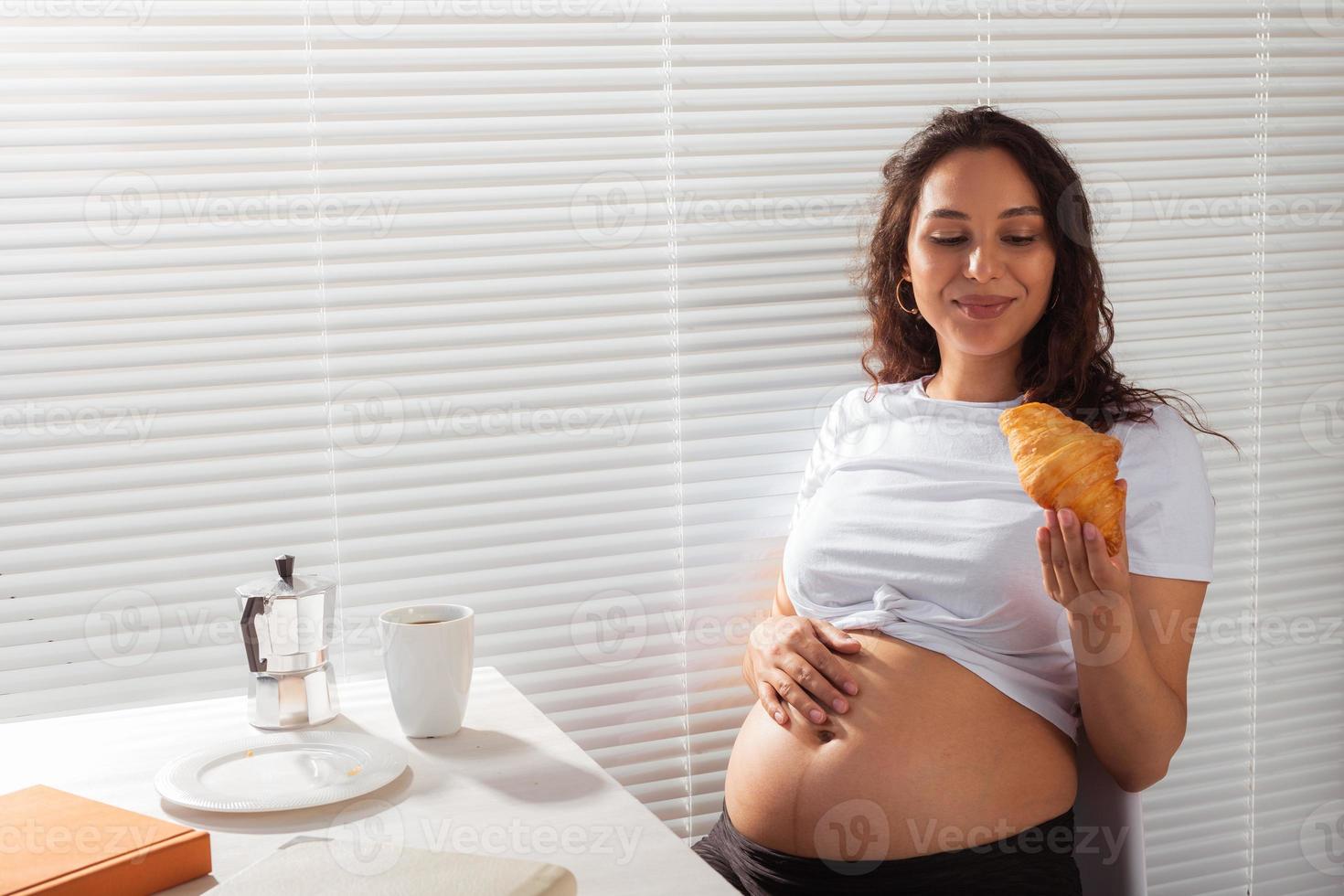 feliz embarazada joven hermosa mujer comiendo croissant durante el desayuno de la mañana. concepto de mañana agradable y actitud positiva durante el embarazo foto