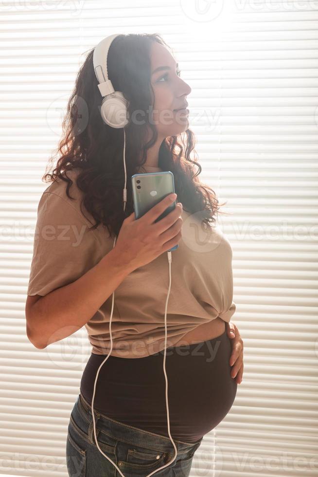 Happy pregnant woman with curly hair listening music in headphones photo