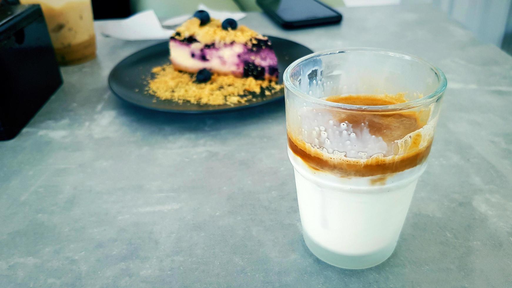 Glass of Dirty coffee with blurred blueberry crumble on black dish or plate and smartphone, phone background on gray or gray concrete table at cafe shop. Food and object with copy space on left. photo