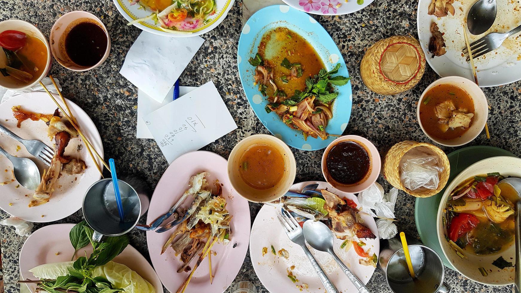 Flat lay of Thai food with spicy papaya salad, white noodles, bone chicken, crab salad, sauce and fresh tomato remain on table after eat lunch at restaurant. Food waste concept. photo