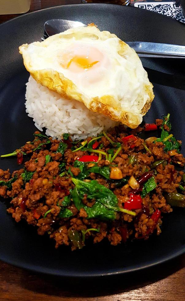 cierre la carne picada salteada picante con albahaca y huevo frito sobre arroz blanco al estilo tailandés en un plato o plato negro en un restaurante local. comida famosa o asiática en tailandia. foto