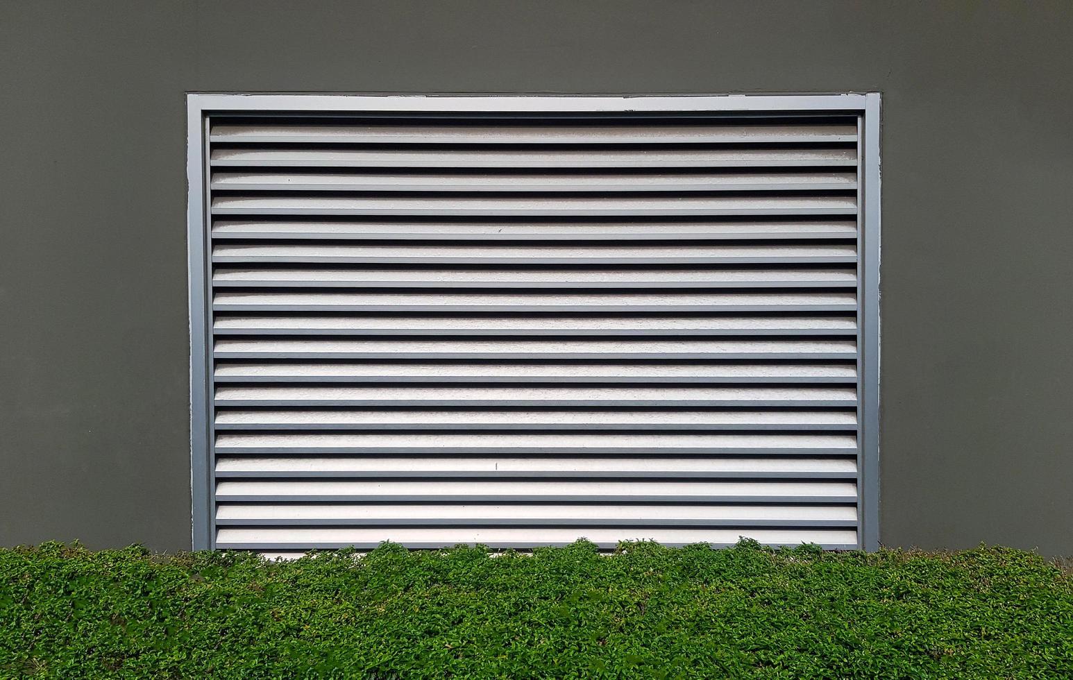 Stainless steel window or door shutter install on gray or grey wall with green bush or tree foreground. Building and Exterior design for access. photo