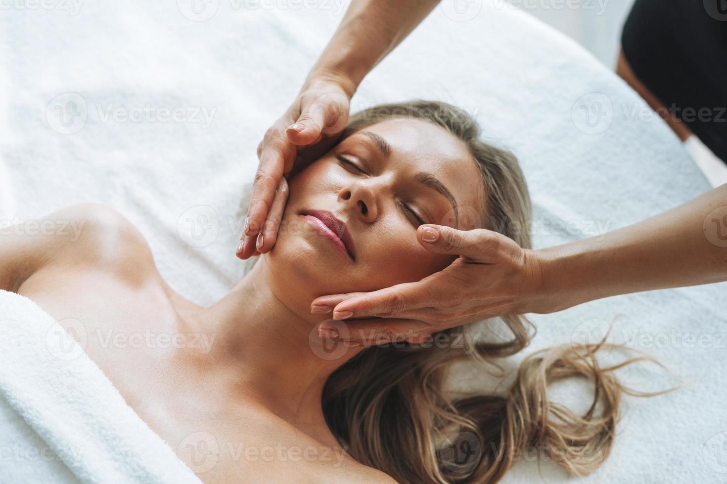 Attractive blonde young woman on couch with white linen enjoys hand facial massage in spa salon, treat yourself photo