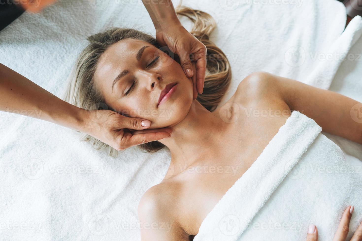 atractiva joven rubia en un sofá con lino blanco disfruta de un masaje facial en el salón de spa, date un capricho foto