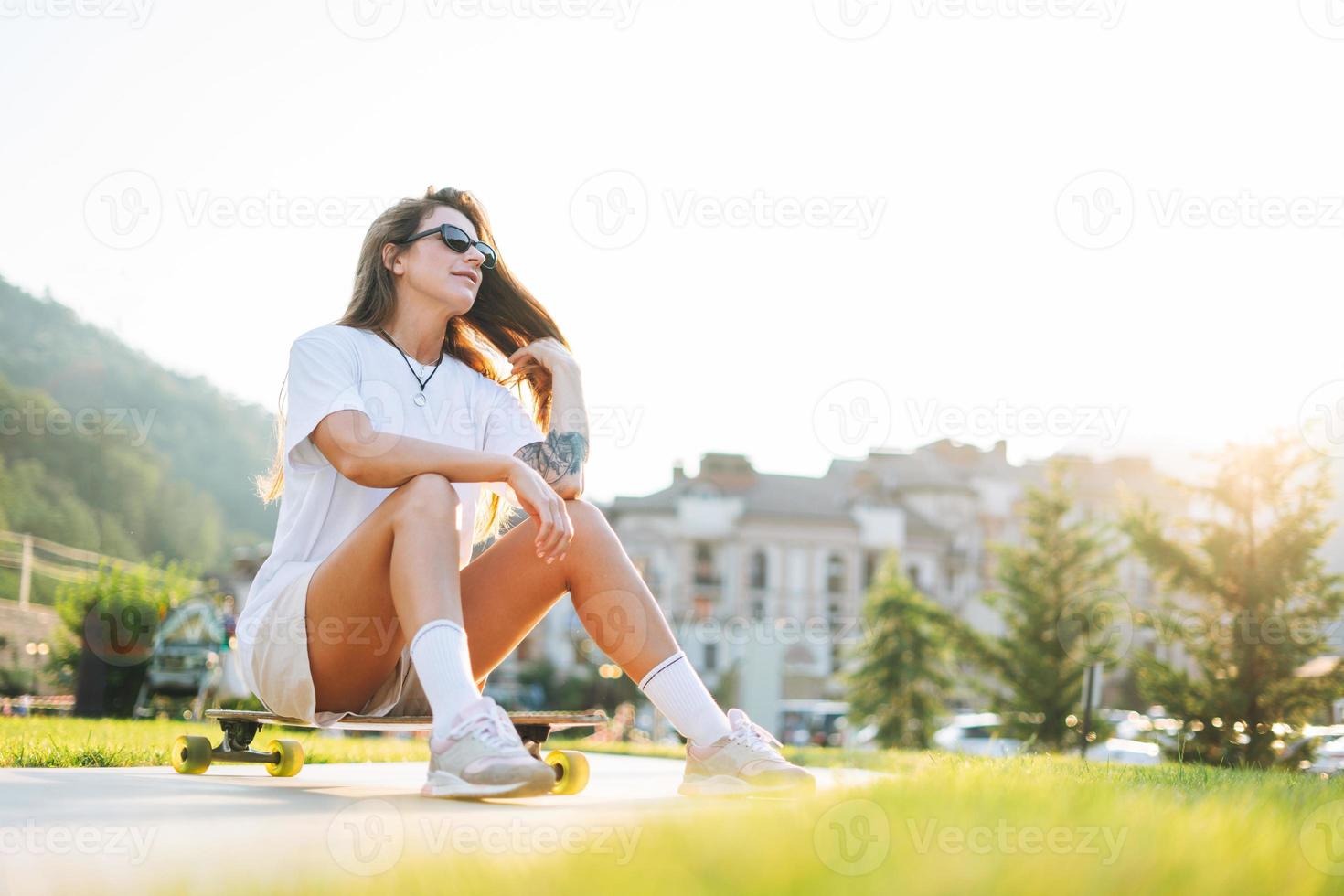 Slim young woman with long blonde hair in light sports clothes sitting with longboard in the outdoor skatepark at the sunset photo