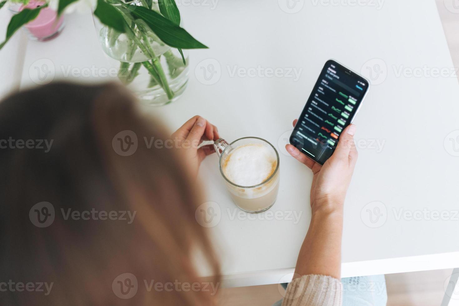 Young woman checking cryptocurrency charts and stock quotes on mobile phone with cup of coffee in the kitchen at home, view from top photo