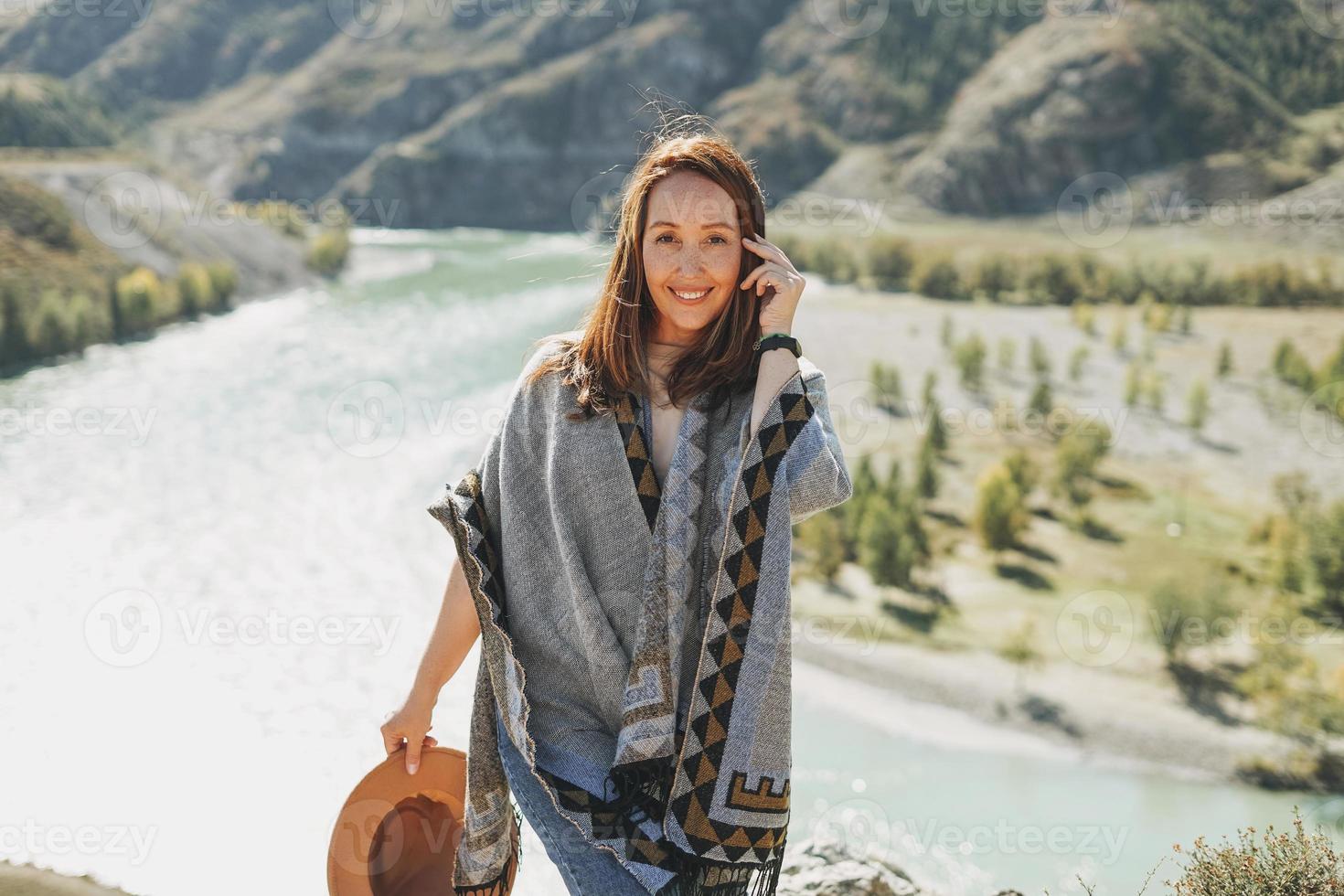 Carefree brunette young woman traveler in poncho on background of the mountain river photo