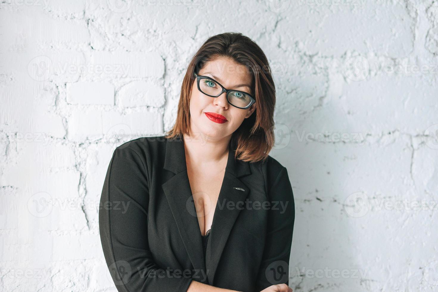 hermosa mujer joven y sonriente mujer de negocios con gafas y chaqueta negra contra la pared blanca foto