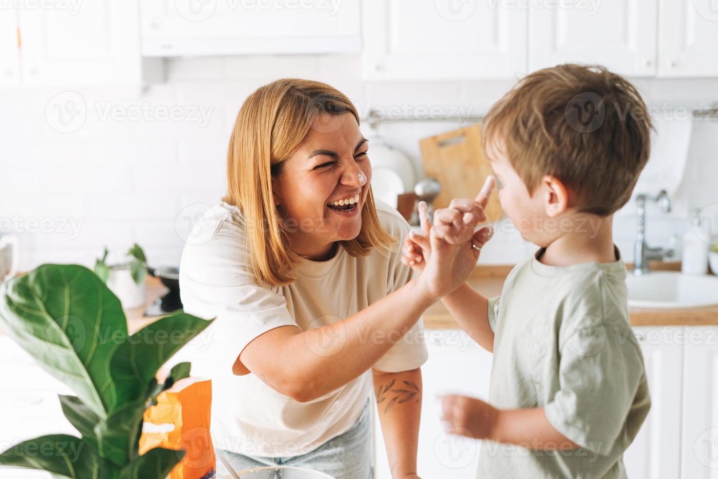 Young woman mother and her toddler boy son have fun while cooking with flour at table in bright kitchen at home photo