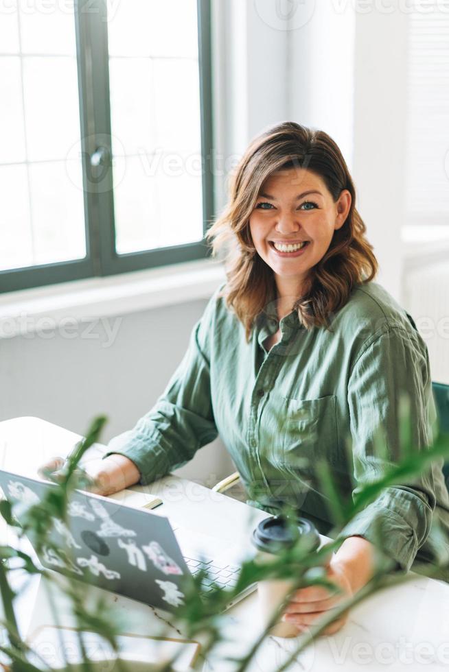 Young smiling brunette woman plus size working at laptop on table with house plant in bright modern office photo