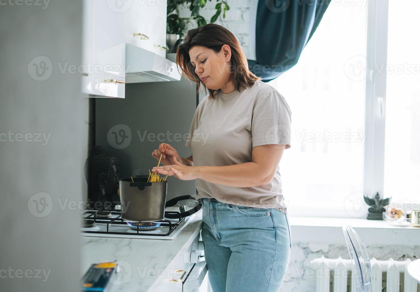 Beautiful brunette young woman plus size body positive cooking pasta in kitchen at home photo