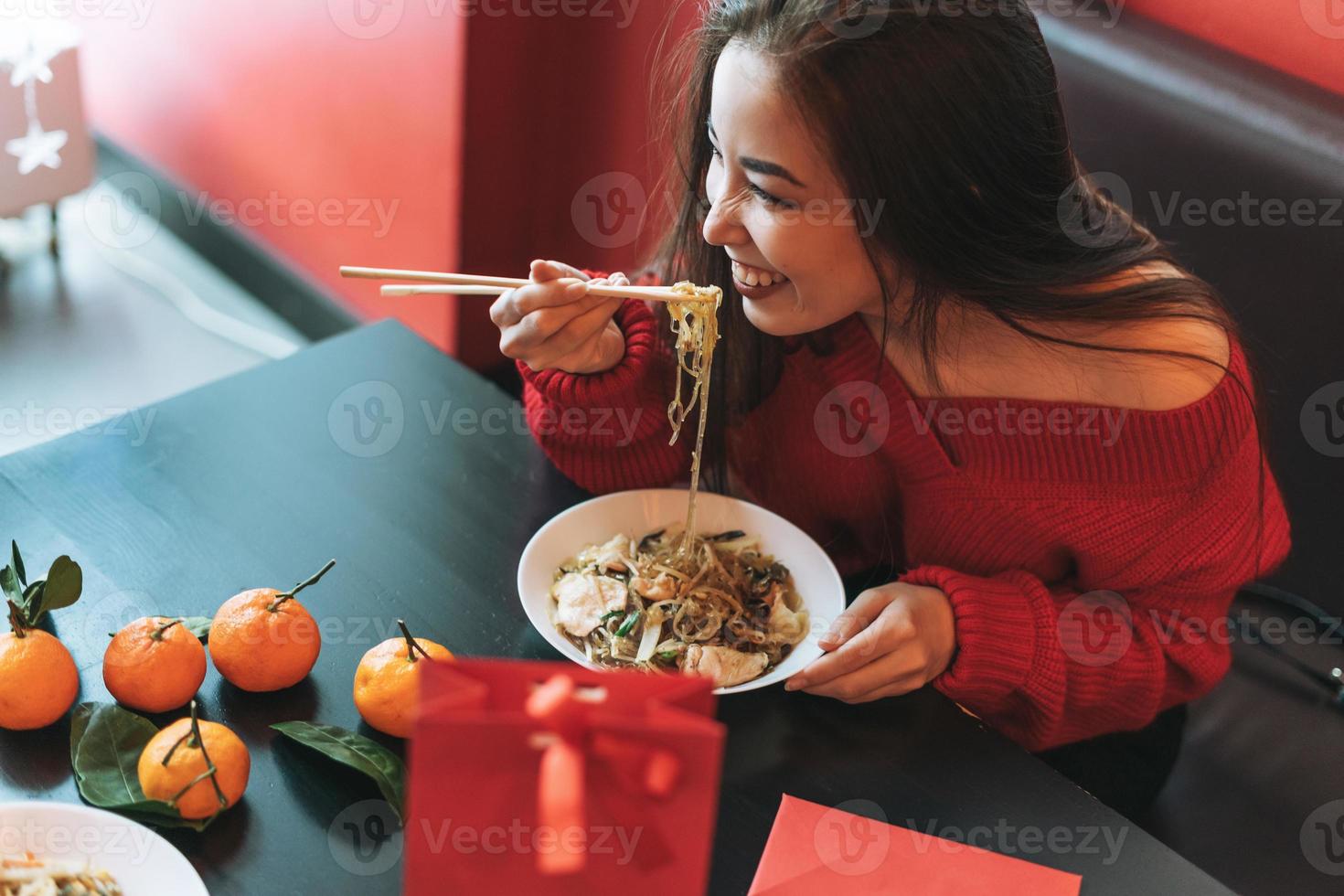 Beautiful smiling young asian woman in red clothes eating noodles with bamboo chopsticks in the chinese vietnamese restaurant photo