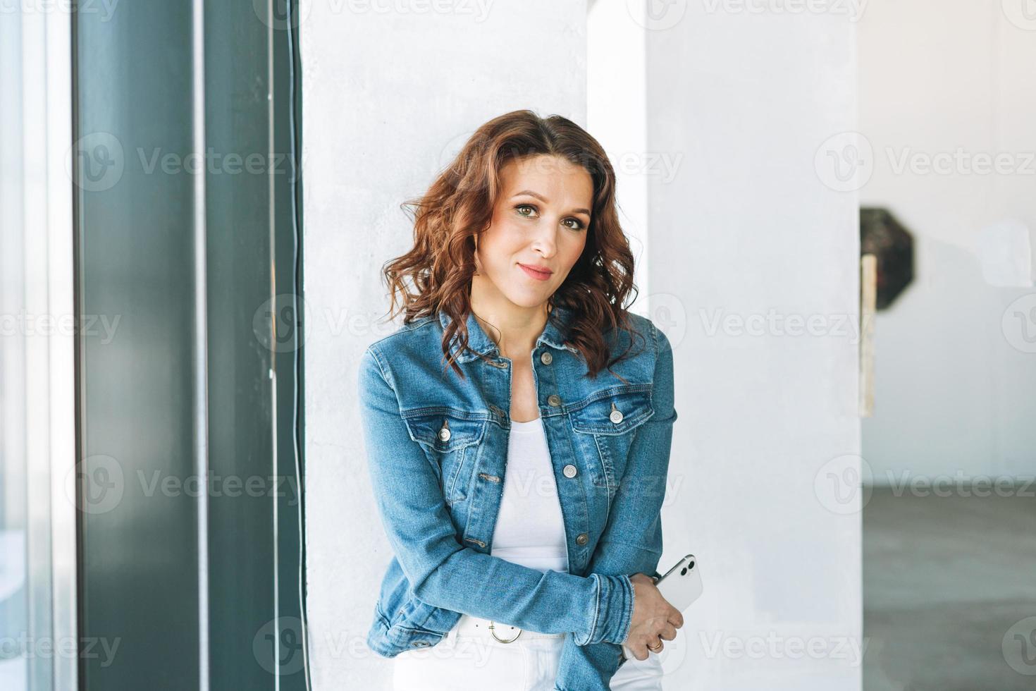 Portrait of smiling charming young woman in jeans jacket with mobile phone near window in modern building photo