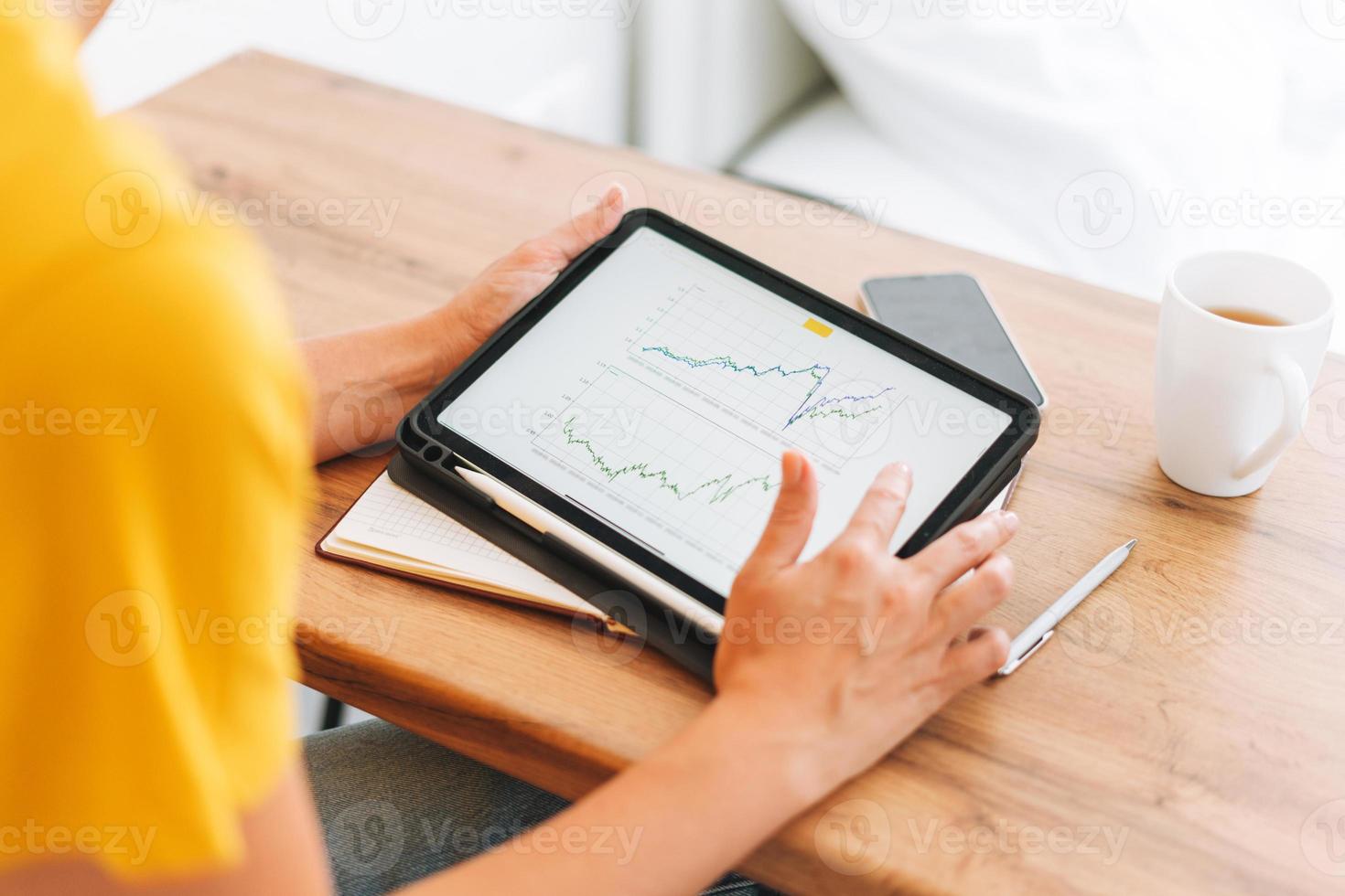 Young blonde woman in yellow t-shirt studies cryptocurrency charts and stock quotes on digital tablet with cup of tea at home photo