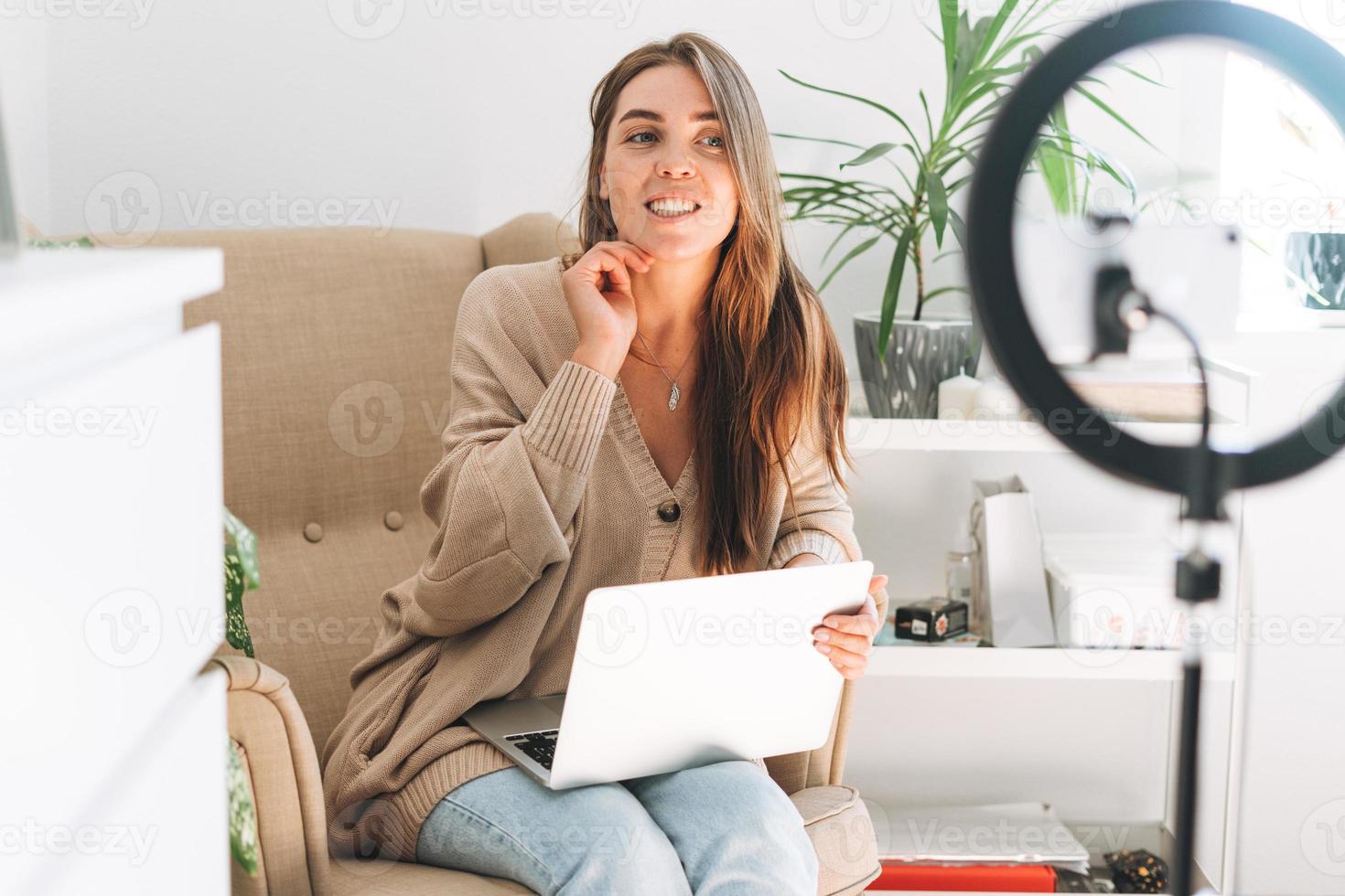 Young pretty woman blogger records video on mobile phone using ring lamp sitting on chair with laptop in room with green plants at home photo