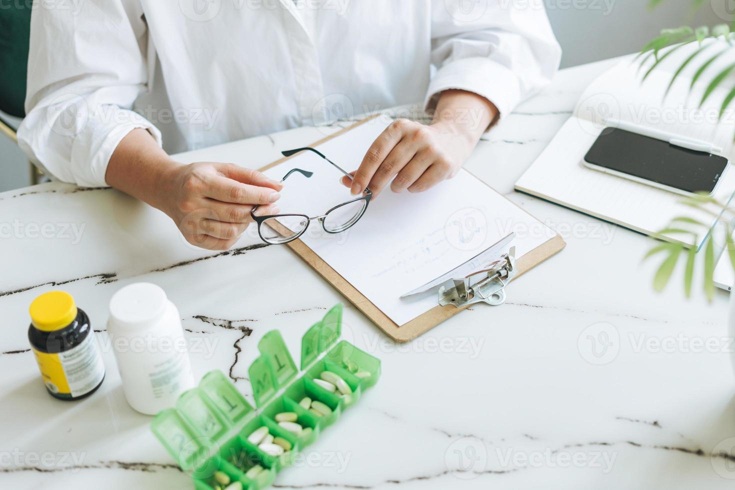 foto recortada de una doctora nutricionista de talla grande con camisa blanca trabajando en la mesa con pastillas y notas diarias en una sala de oficina moderna y luminosa