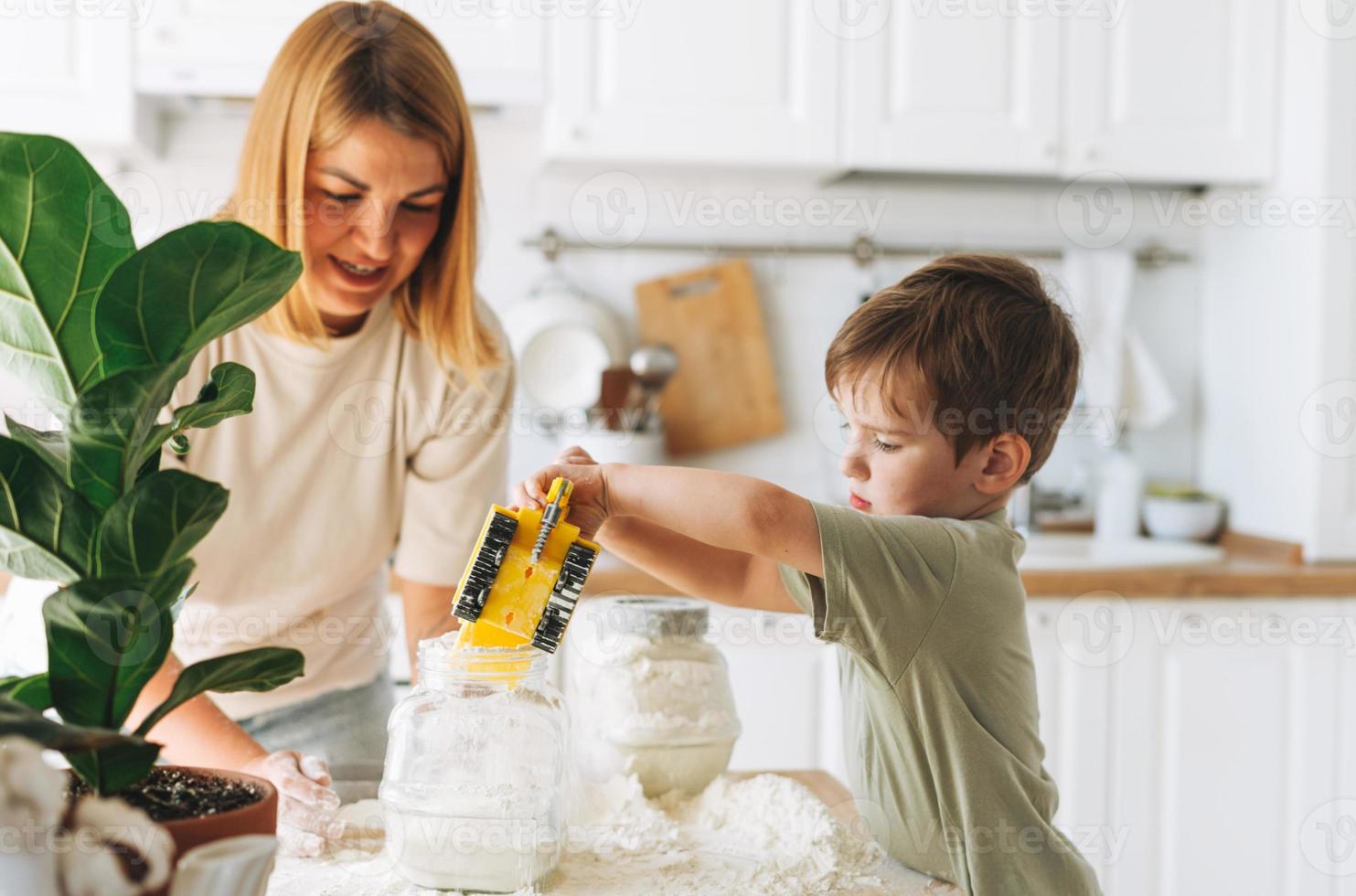 Young woman mother and her toddler boy son have fun while cooking with flour at the table in kitchen at home. Boy playing with car toys photo