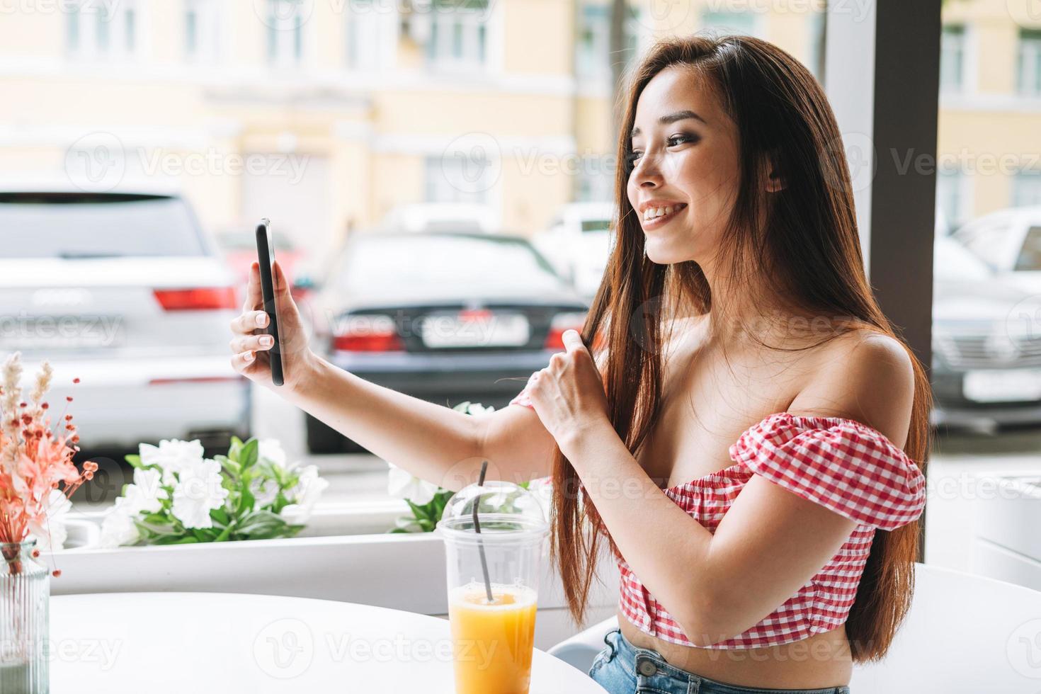 sonriente hermosa joven asiática con el pelo largo rastrillando selfie en el teléfono móvil en la terraza del café de verano. chica vestida de rojo de vacaciones en la ciudad turística foto