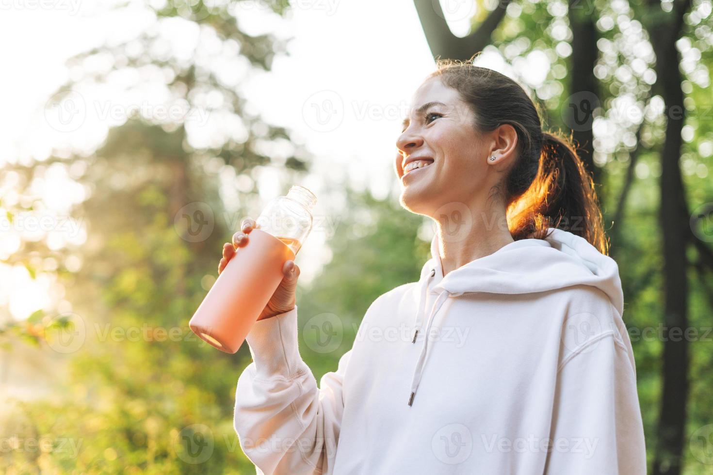 Young slim woman brunette in sport clothes running and drinking water at forest on golden hour sunrise time. Health and wellness, fitness lifestyle photo