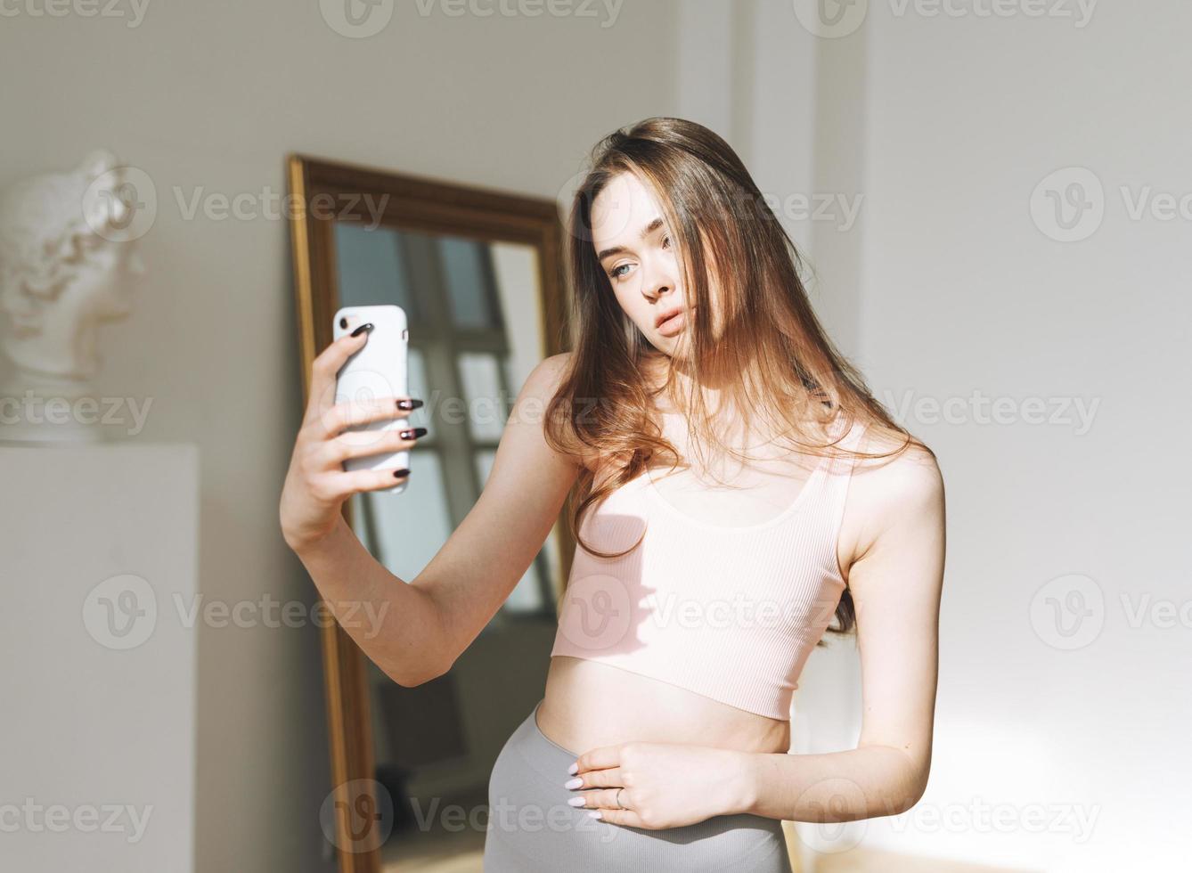 Beautiful young woman with long hair in sport wear taking selfie using mobile in studio with hard sun light photo