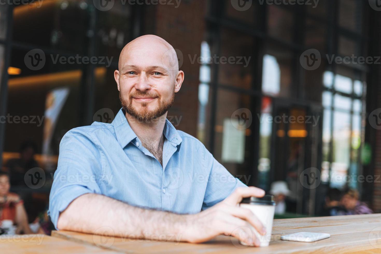 Adult bald smiling attractive man forty years with beard in blue shirt businessman using mobile phone with paper cup of coffee at cafe photo