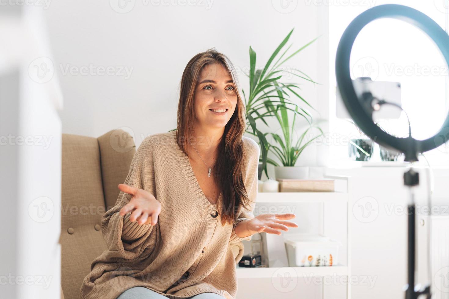 joven bloguera de mujer bonita en cardigan acogedor graba video en el teléfono móvil usando una lámpara de anillo sentada en una silla en el hogar foto