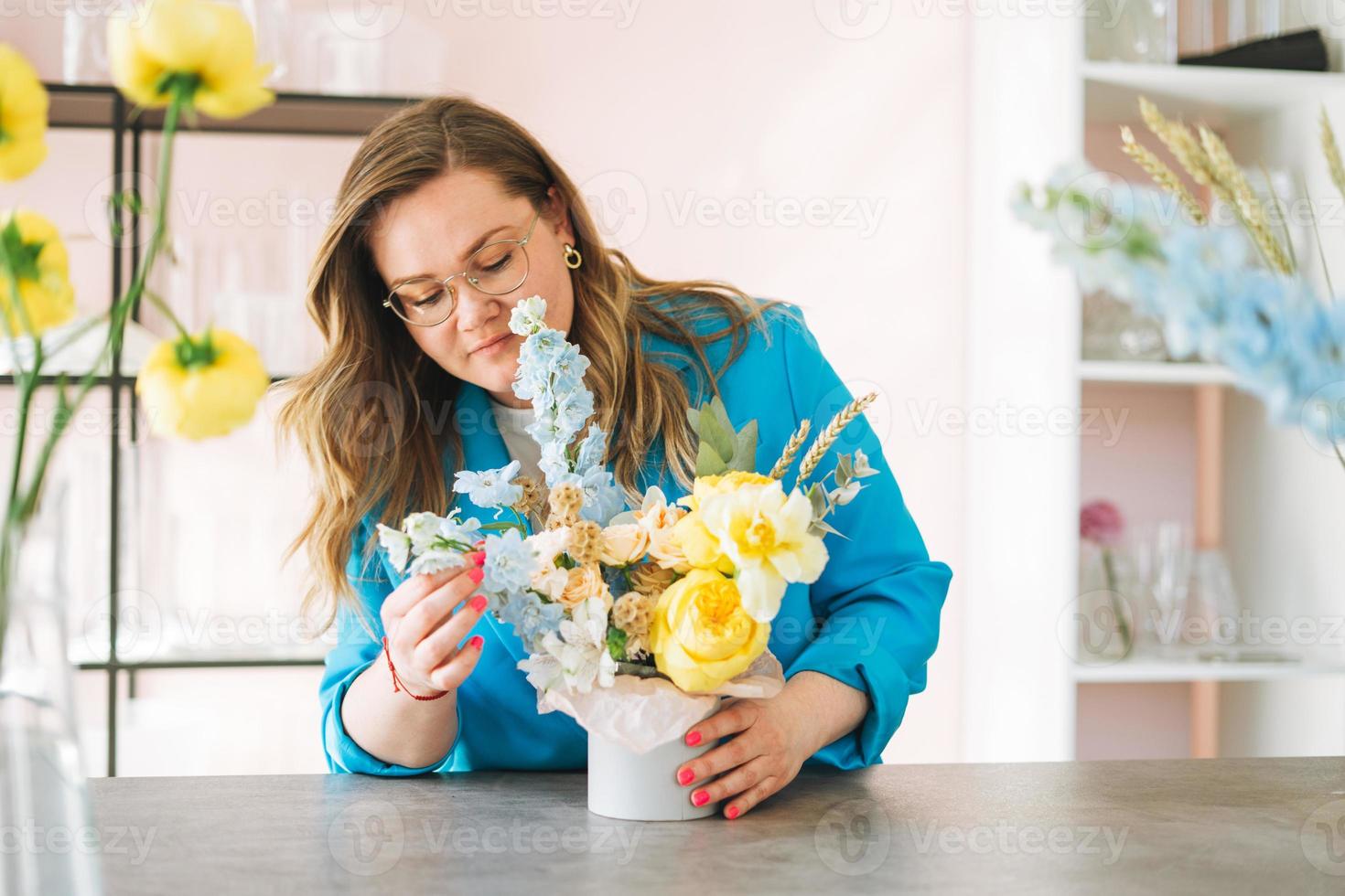 Young woman florist in bright blue suit and eye glasses with bouquet of flowers in box in flower shop, small local business owner. Young stylish success millennial woman on creative work photo