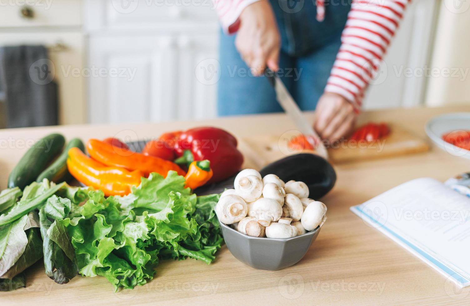 Fresh vegetables green salad, peppers, mushrooms and eggplant on kitchen table. Young woman plus size body positive in red longsleeve cooking using book with recipes on kitchen photo