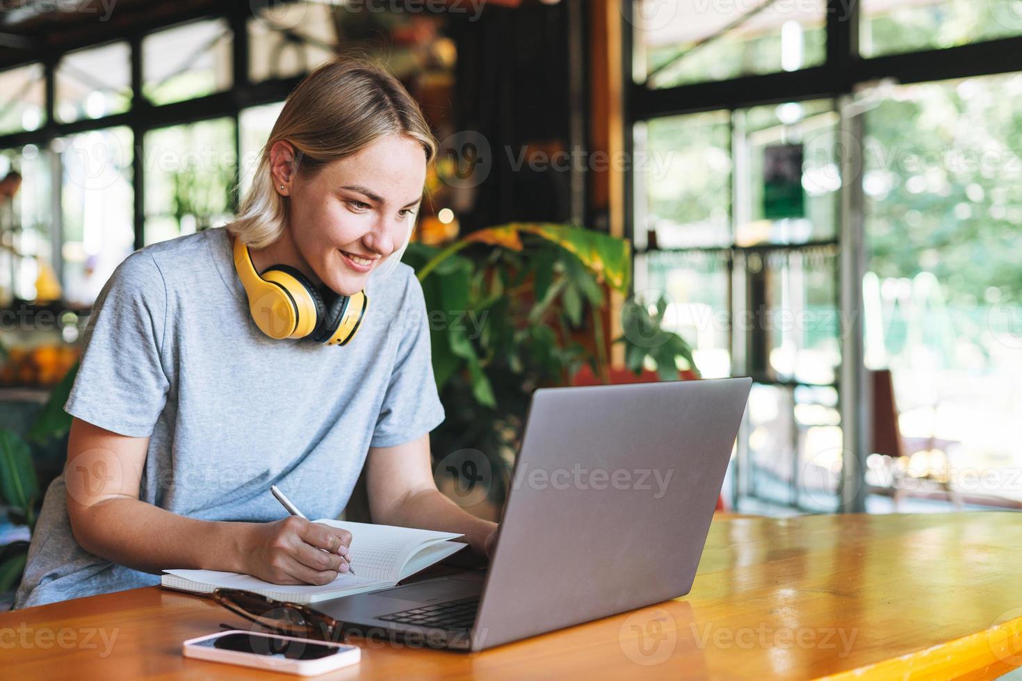 joven mujer rubia sonriente independiente con auriculares amarillos trabajando en una laptop en la mesa del café foto