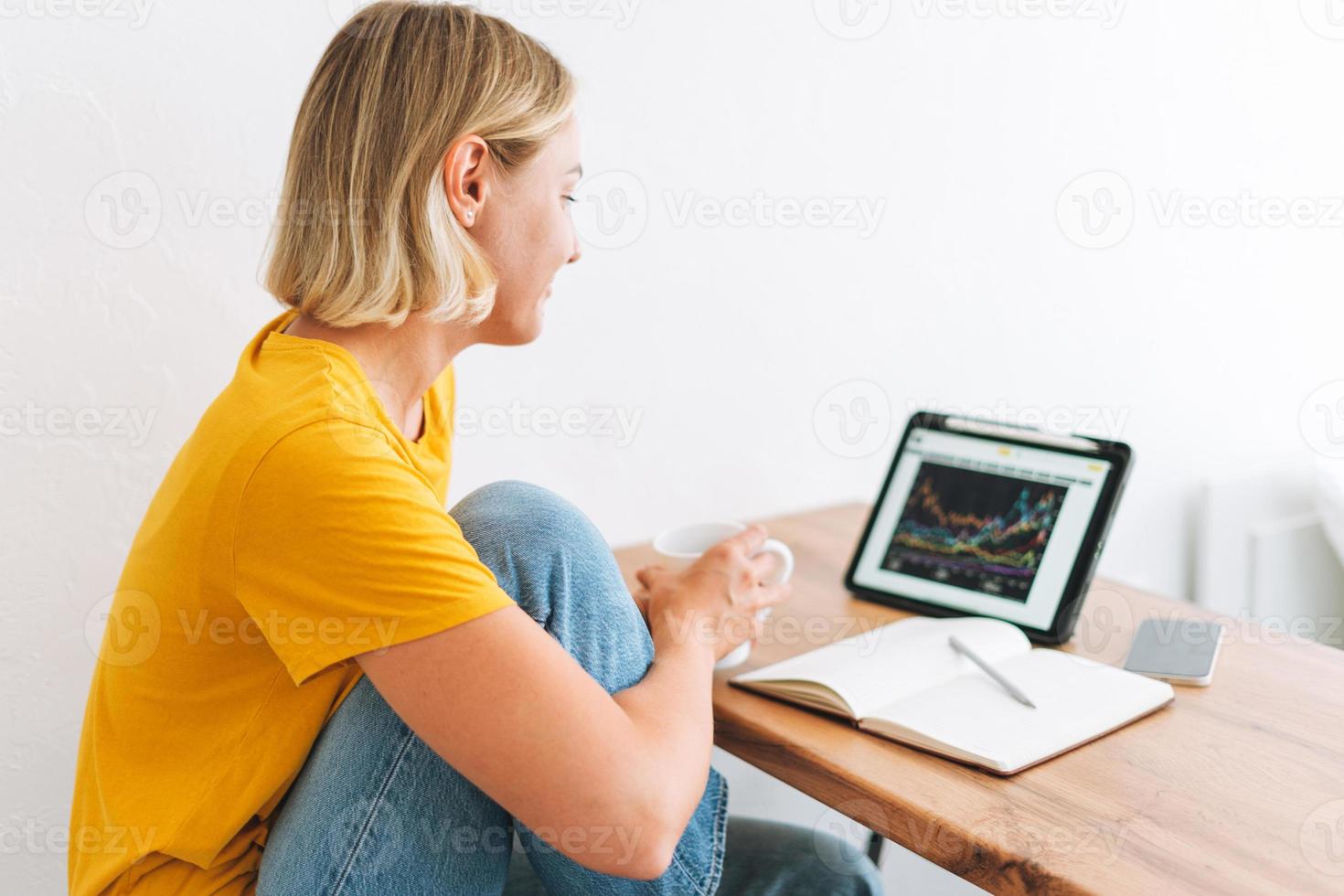 Young blonde woman in yellow t-shirt studies cryptocurrency charts and stock quotes on digital tablet with cup of tea at the home photo