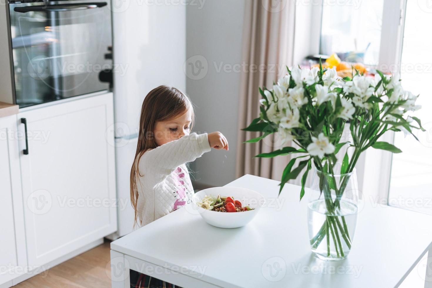 Pretty little girl with long hair sprinkles salt on vegetable salad in kitchen with bright interior at home photo