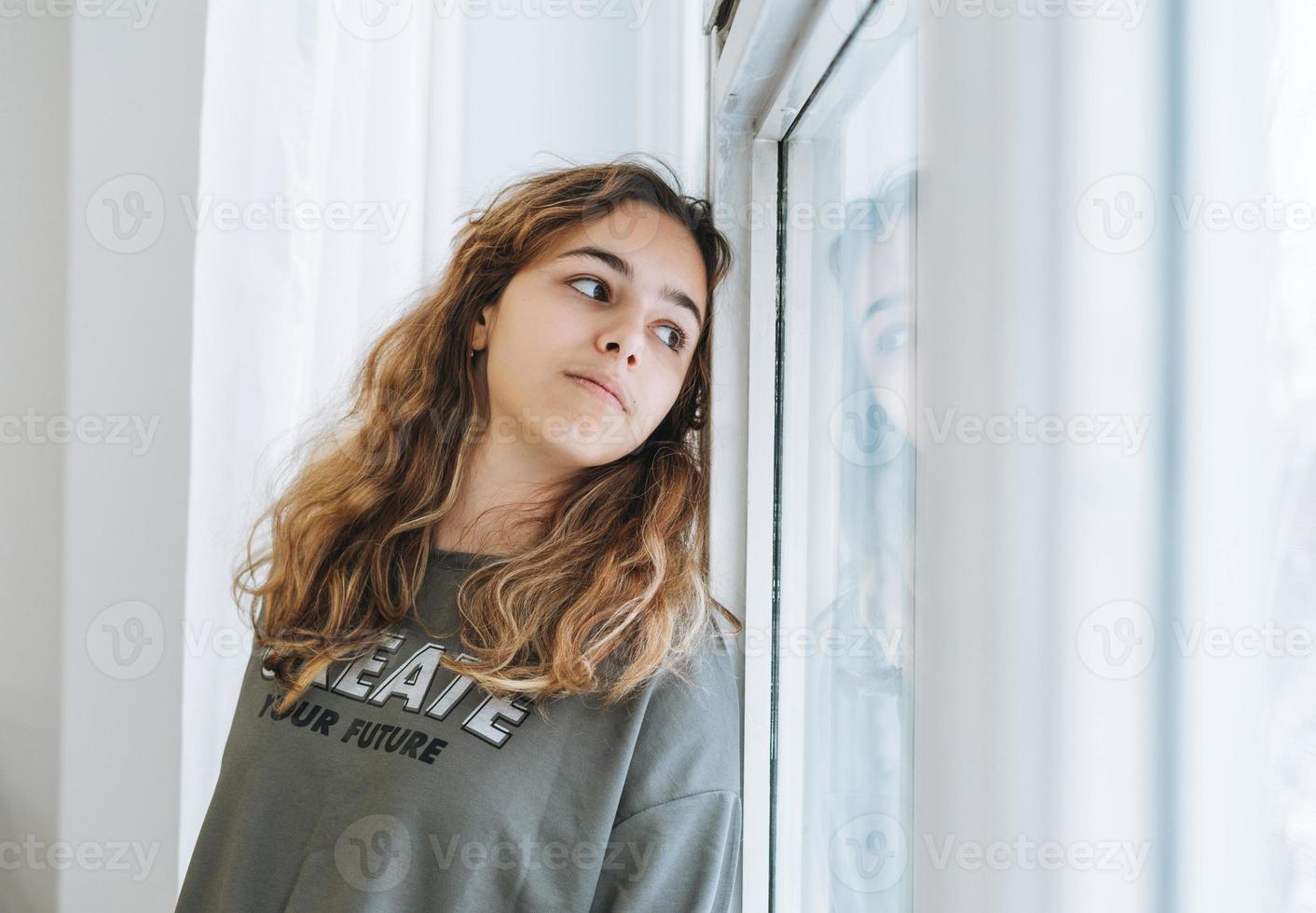 Beautiful sad unhappy teenager girl with curly hair sitting on the window sill photo